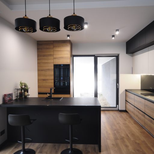 Interior of a modern kitchen, open to the living room, The kitchen has a black half-table with hockers. Cabinet fronts are imitation wood, white and grey.
