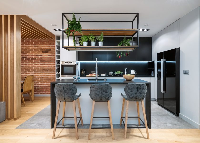 Modern kitchen open to the living room, in the foreground there is an island with hockers, an industrial flowerbed hangs over the island. On the right side there is a light development with a two-door fridge. Worktop illuminated with led strip, upper cabinets in dark colour.