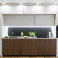 A modern kitchen open to the living room. Lower cabinets of dark wood panel, on both sides high pillars of light panel, in the left one there is a refrigerator, on the right there is a pillar with an oven. Upper cabinets made of light board. The worktop is illuminated by a led strip. The whole unit is illuminated by spotlights.