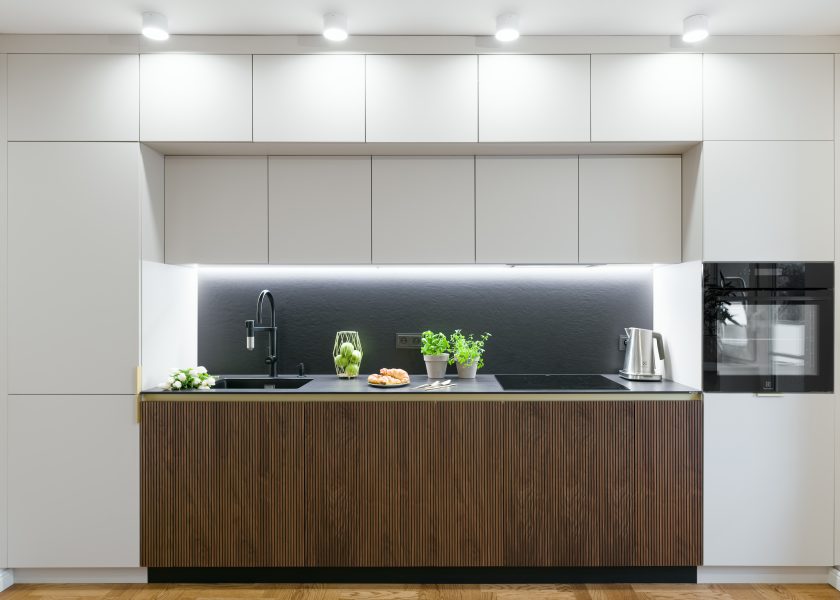 A modern kitchen open to the living room. Lower cabinets of dark wood panel, on both sides high pillars of light panel, in the left one there is a refrigerator, on the right there is a pillar with an oven. Upper cabinets made of light board. The worktop is illuminated by a led strip. The whole unit is illuminated by spotlights.